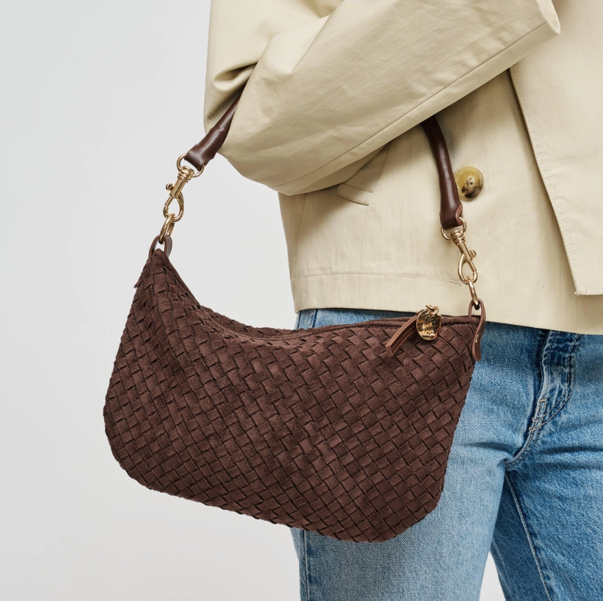 A brown woven leather shoulder bag with gold-tone hardware, shown being worn over the shoulder of a person wearing a beige blazer and blue jeans against a light gray background.