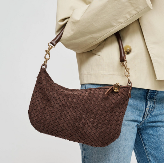 A brown woven leather shoulder bag with gold-tone hardware, shown being worn over the shoulder of a person wearing a beige blazer and blue jeans against a light gray background.