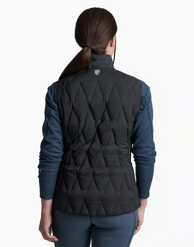 Rear view of a woman wearing a black Kuhl Firestorm diamond-quilted puffer vest over a dark blue long-sleeve shirt and dark gray leggings, against a plain white background.