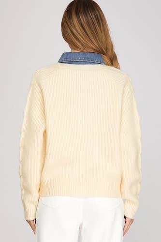 A woman viewed from the back wearing a cream ribbed knit sweater with a blue denim collar, paired with white pants, against a light gray studio background.