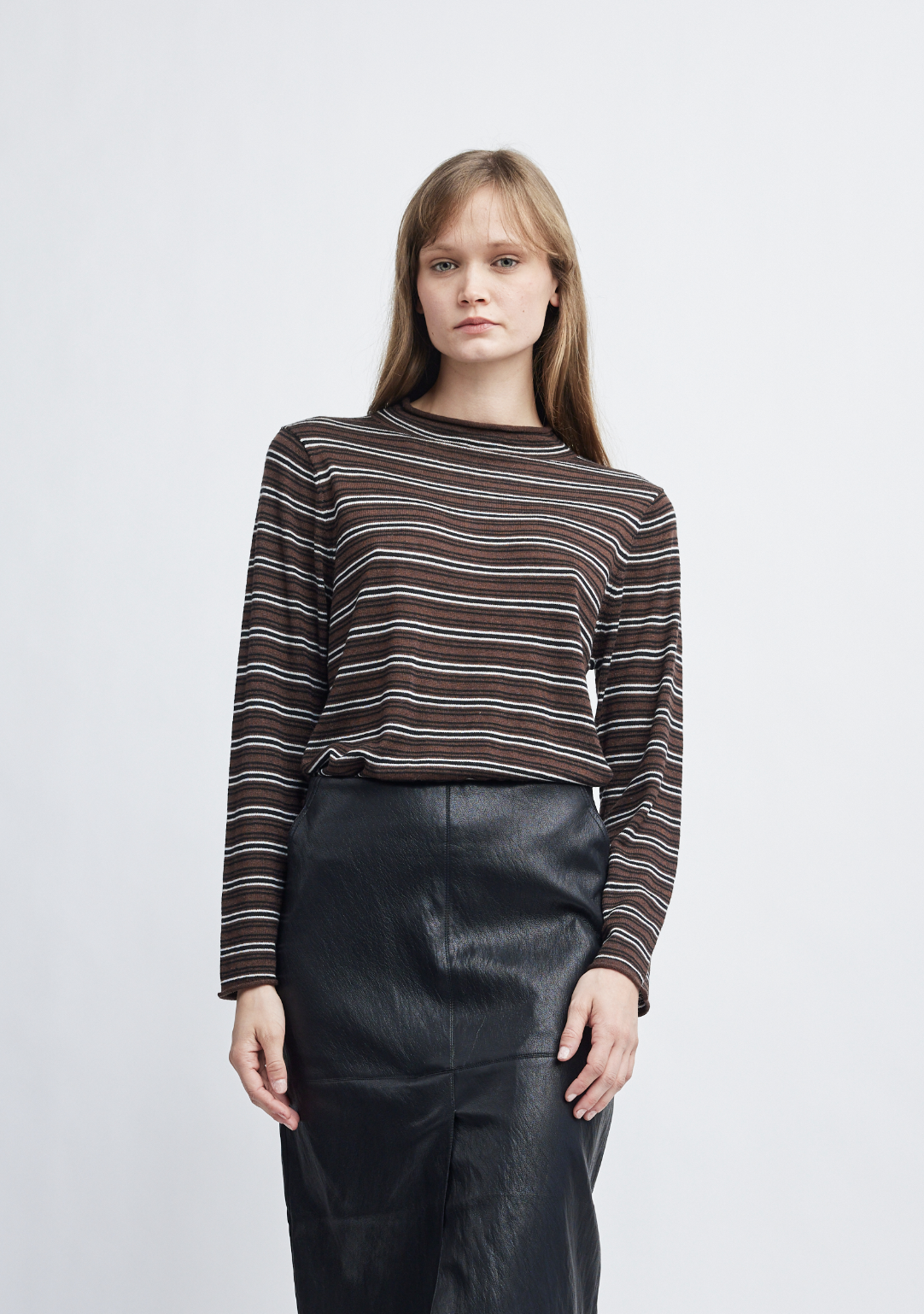 A model wearing a brown and white striped long-sleeve knit top with black leather pants, standing against a light gray studio background.