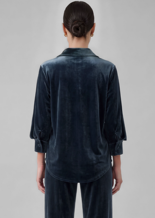 A woman viewed from the back wearing a dark navy velvet long-sleeve collared shirt and matching dark pants against a light gray studio background.