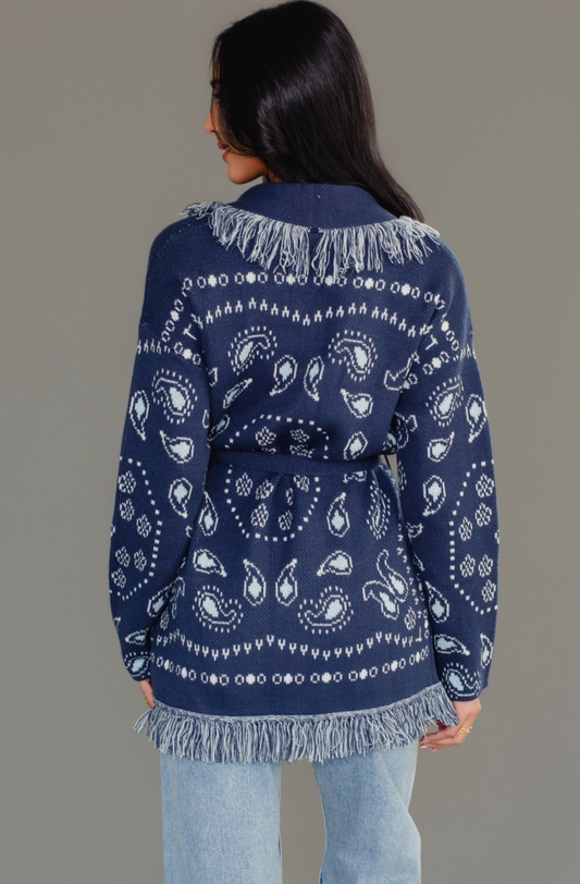 A woman shown from the back wearing a navy blue knit cardigan with white paisley and geometric bandana-style patterns and fringed trim, paired with light blue jeans against a plain gray studio background.