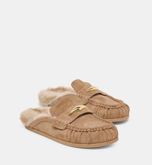 A pair of Dolce Vita Jodero tan suede slip-on mule flats with a soft cream shearling lining and a small gold-tone metal accent on the upper, shown on a light gray studio background.