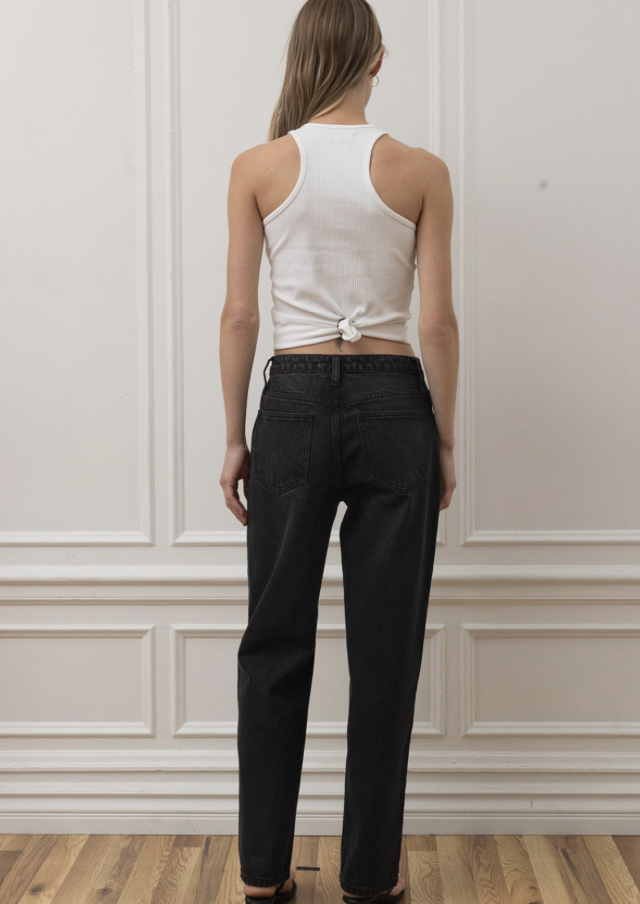 A woman shown from the back wearing a white ribbed tank top with a knot detail at the hem and black straight-leg jeans, standing against a white paneled wall.