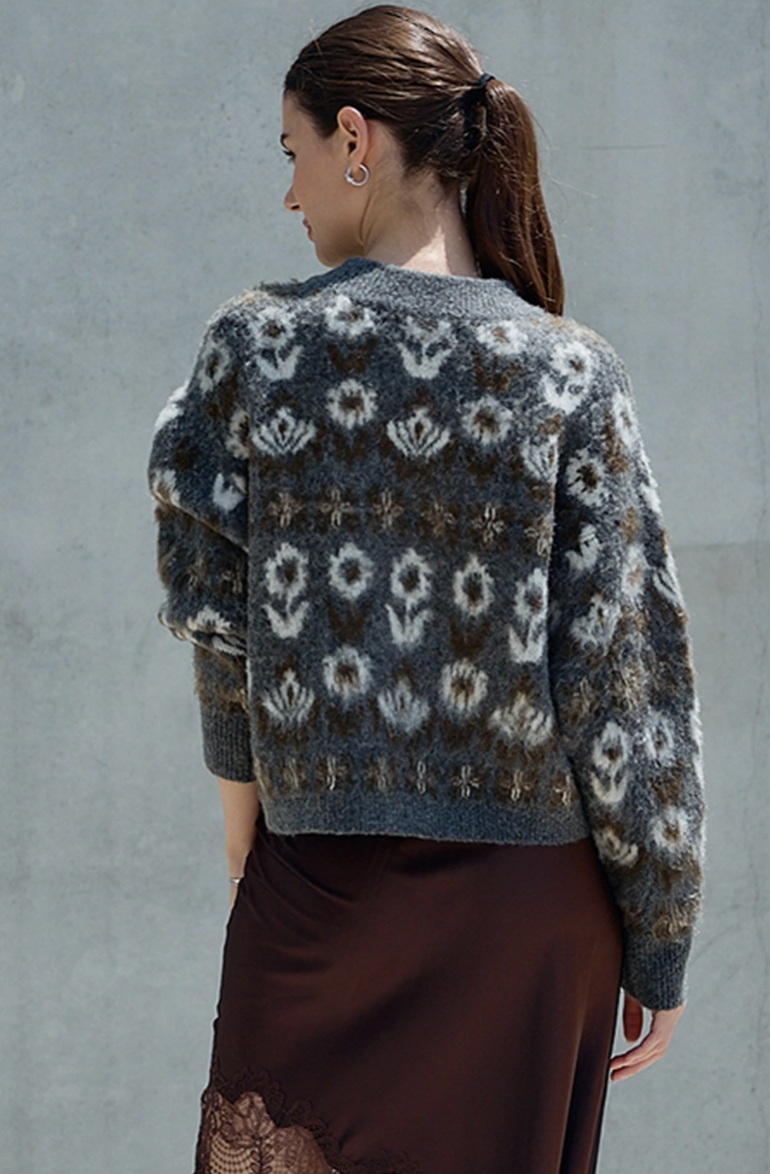 A woman shown from the back wearing a fuzzy knit cardigan with a gray, brown, and off-white floral pattern, paired with a dark brown skirt, set against a textured gray wall.