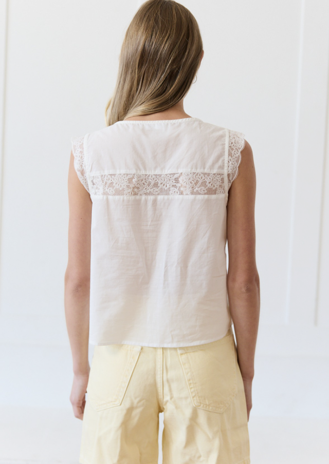 A woman viewed from behind wearing a sleeveless cream-colored top with a lace band across the upper back and pale yellow pants, set against a bright white background.