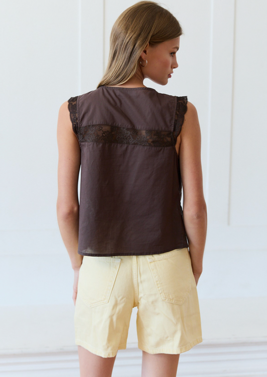 A woman shown from the back wearing a sleeveless dark brown top with a sheer lace band across the upper back and light yellow shorts, set against a bright white paneled wall.