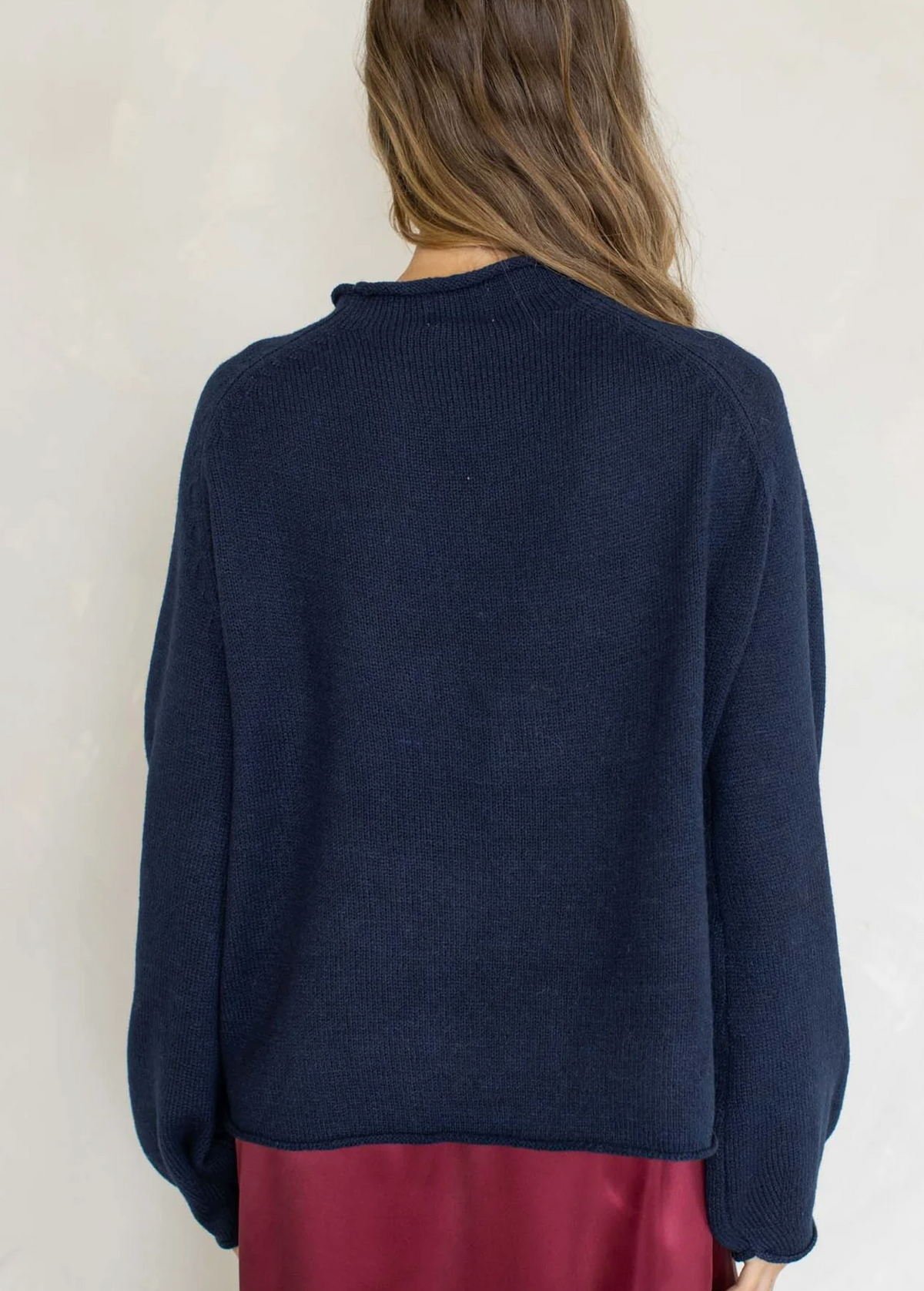 A woman viewed from the back wearing a dark navy blue knit sweater and a burgundy skirt, set against a plain off-white wall.
