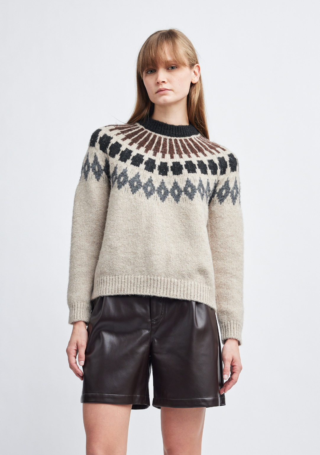 A woman modeling a beige knit sweater with a dark brown and gray geometric Fair Isle yoke pattern, paired with dark brown faux leather shorts, against a light gray studio background.