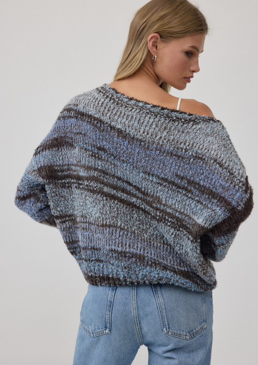A woman wearing a chunky knit off-the-shoulder sweater with horizontal marled stripes in shades of blue, gray, and brown, paired with light-wash jeans against a plain light gray background.