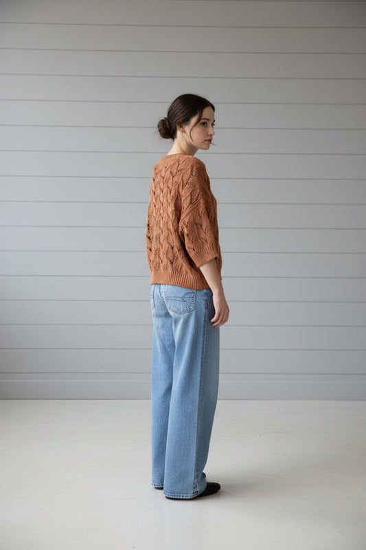 A person viewed from behind wearing a loose, textured knit sweater in a warm terracotta or rust color, paired with light blue jeans, standing outdoors next to a dark door and brick wall with metal mailboxes.