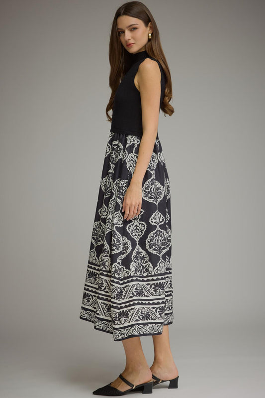 A woman modeling a sleeveless black midi dress with a black and white floral ikat patterned skirt, paired with black mule heels, against a solid gray studio background.