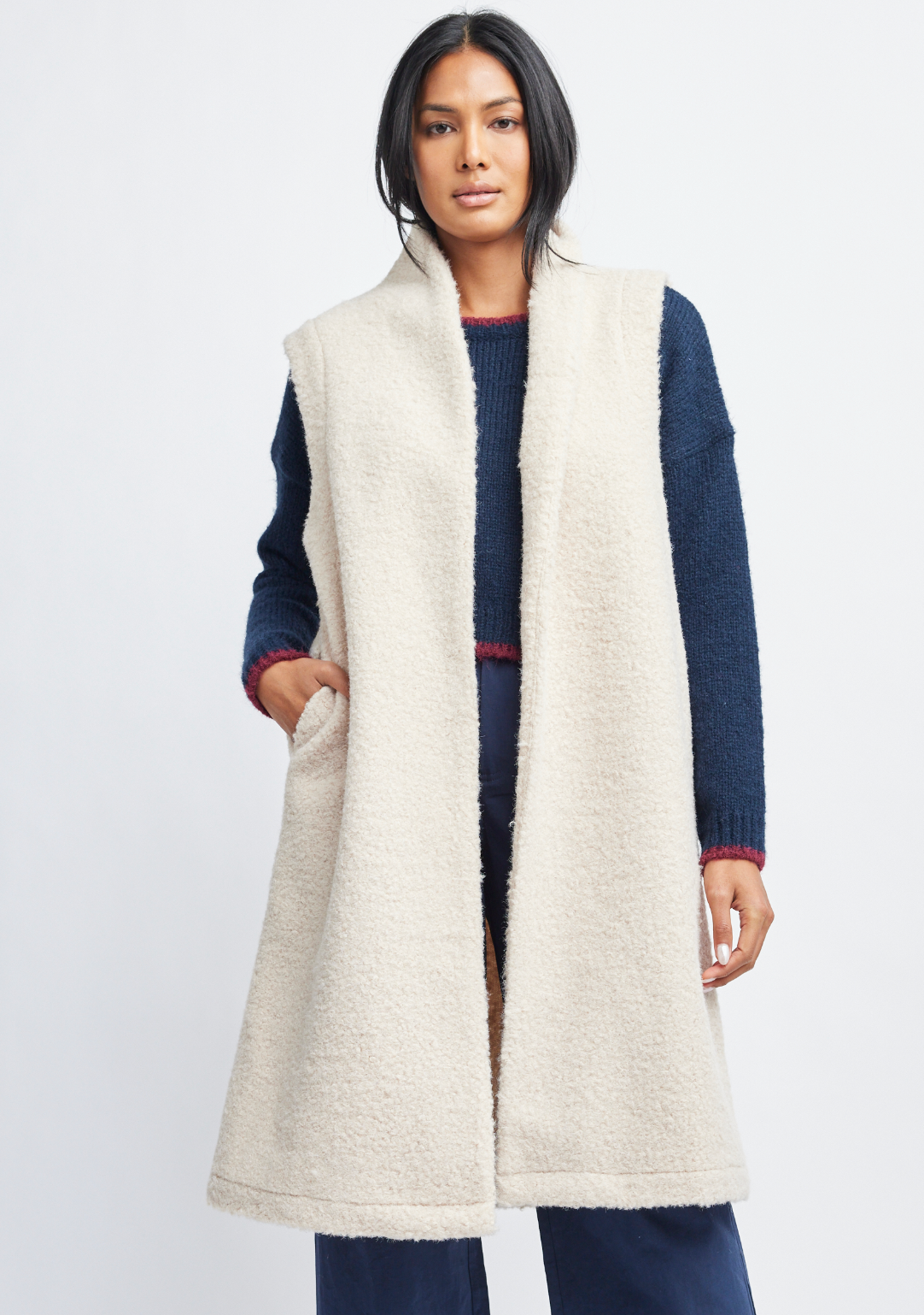 A woman modeling a long cream sherpa fleece vest over a navy blue knit sweater and navy pants, photographed against a clean light gray studio background.