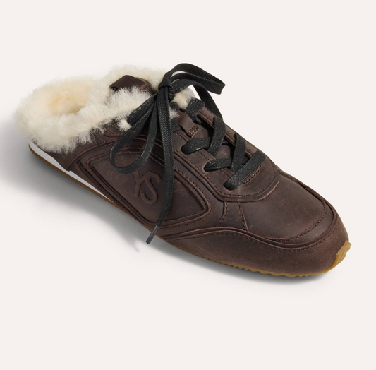 A Yosi Samra dark brown leather slip-on sneaker with a fluffy cream shearling lining and black laces, shown from a three-quarters angle on a light beige background.
