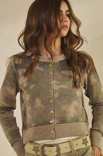 A woman wearing a lightweight cropped button-front cardigan with a muted camouflage pattern in olive green, beige, and brown tones, paired with a light belt, against a plain beige background.