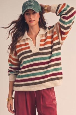A woman wearing a cozy, long-sleeve knit polo sweater with horizontal stripes in cream, orange, brown, green, and burgundy, paired with rust red pants and a green baseball cap.