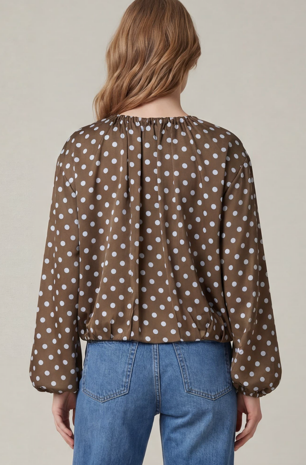 A woman viewed from the back wearing a brown long-sleeve blouse with white polka dots and blue jeans, against a plain light beige studio background.
