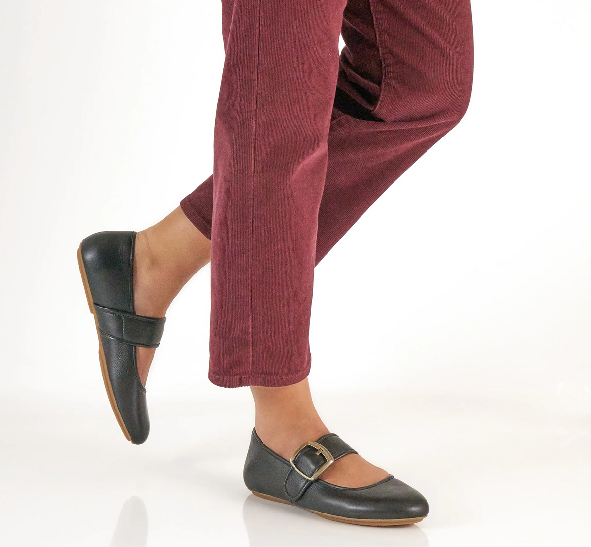 A person wearing burgundy corduroy pants and Dansko Matilda black leather Mary Jane flats with gold-tone buckle straps, shown from the knees down against a clean white studio background.