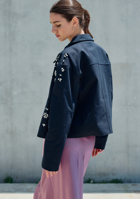 A woman wearing a dark navy cropped jacket with white floral embroidery on the chest and sleeves, paired with lavender trousers, standing against a textured gray concrete wall.