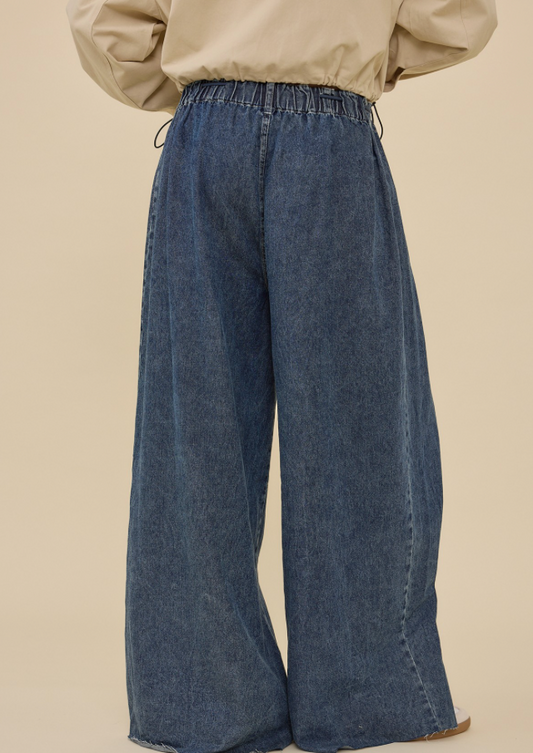 A person wearing wide-leg blue denim jeans with an elastic waistband and drawstrings, paired with a beige top, shown from the back against a plain beige studio background.