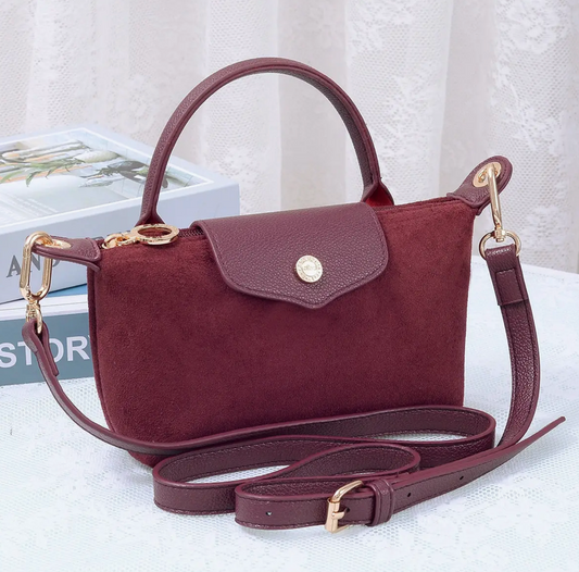 A burgundy suede handbag with a top handle, flap closure, and matching detachable shoulder strap, styled on a white textured surface with a book in the background.