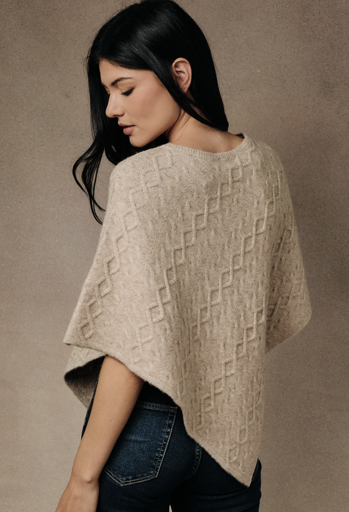 A woman wearing a beige textured knit poncho sweater and dark blue jeans, photographed from a side-back angle against a neutral textured studio background.