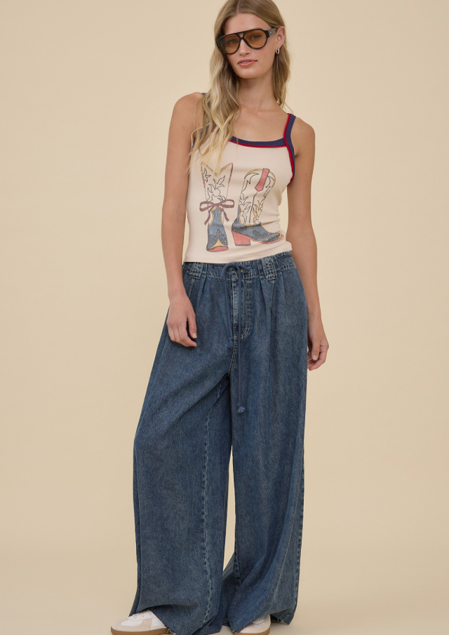 A woman modeling a sleeveless cream tank top with a colorful graphic print and blue denim wide-leg pants, standing against a solid beige background.