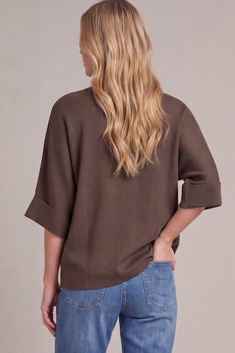 A woman shown from the back wearing a loose-fitting brown top with rolled three-quarter sleeves and blue jeans, against a plain light gray studio background.