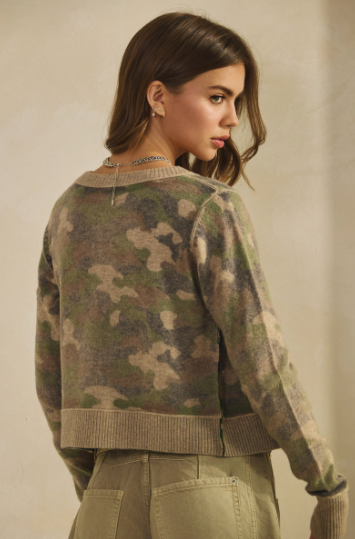 A woman shown from the back wearing a cropped long-sleeve knit sweater with an olive green, brown, and beige camouflage pattern, paired with khaki pants, against a neutral beige background.