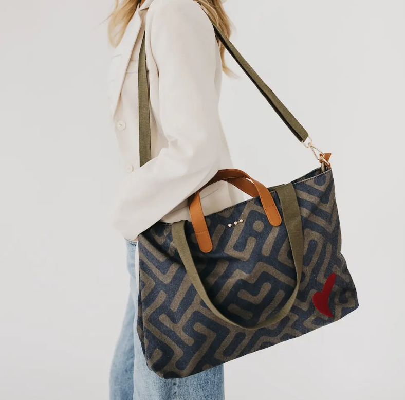 A person carrying a large patterned canvas tote bag with olive green handles and strap, featuring a small red heart detail on the side, against a plain white background.
