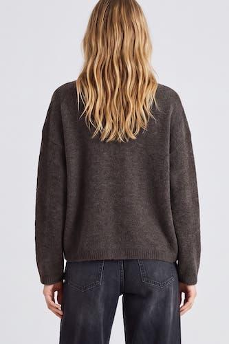 A woman viewed from the back wearing a dark brown knit sweater and dark jeans against a light gray studio background.