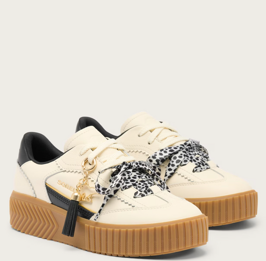 A pair of Sorel low-top platform sneakers in off-white leather with black leather heel tabs, brown gum rubber soles, and decorative black-and-white leopard-print ribbon bows on the laces. A gold-tone tassel charm with a black tassel and a gold brand name plate is attached to the side of the front shoe.