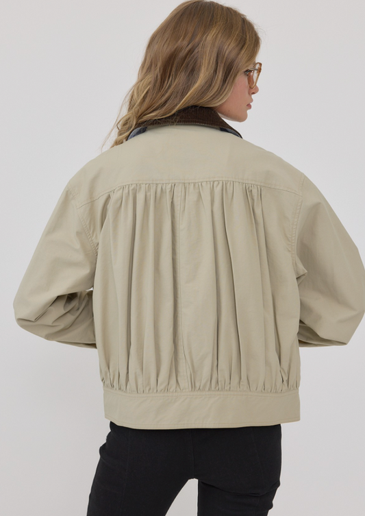 A woman shown from the back wearing a light beige cropped jacket with gathered pleating across the back, black pants, and glasses against a plain light gray studio background.