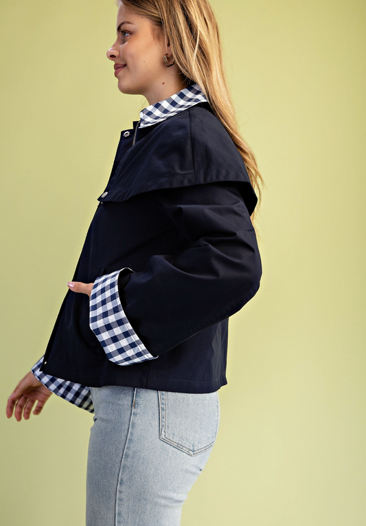 A woman shown in side profile wearing a dark navy jacket with a contrasting blue and white gingham collar and cuffs, paired with light-wash jeans, against a solid pale yellow-green studio background.