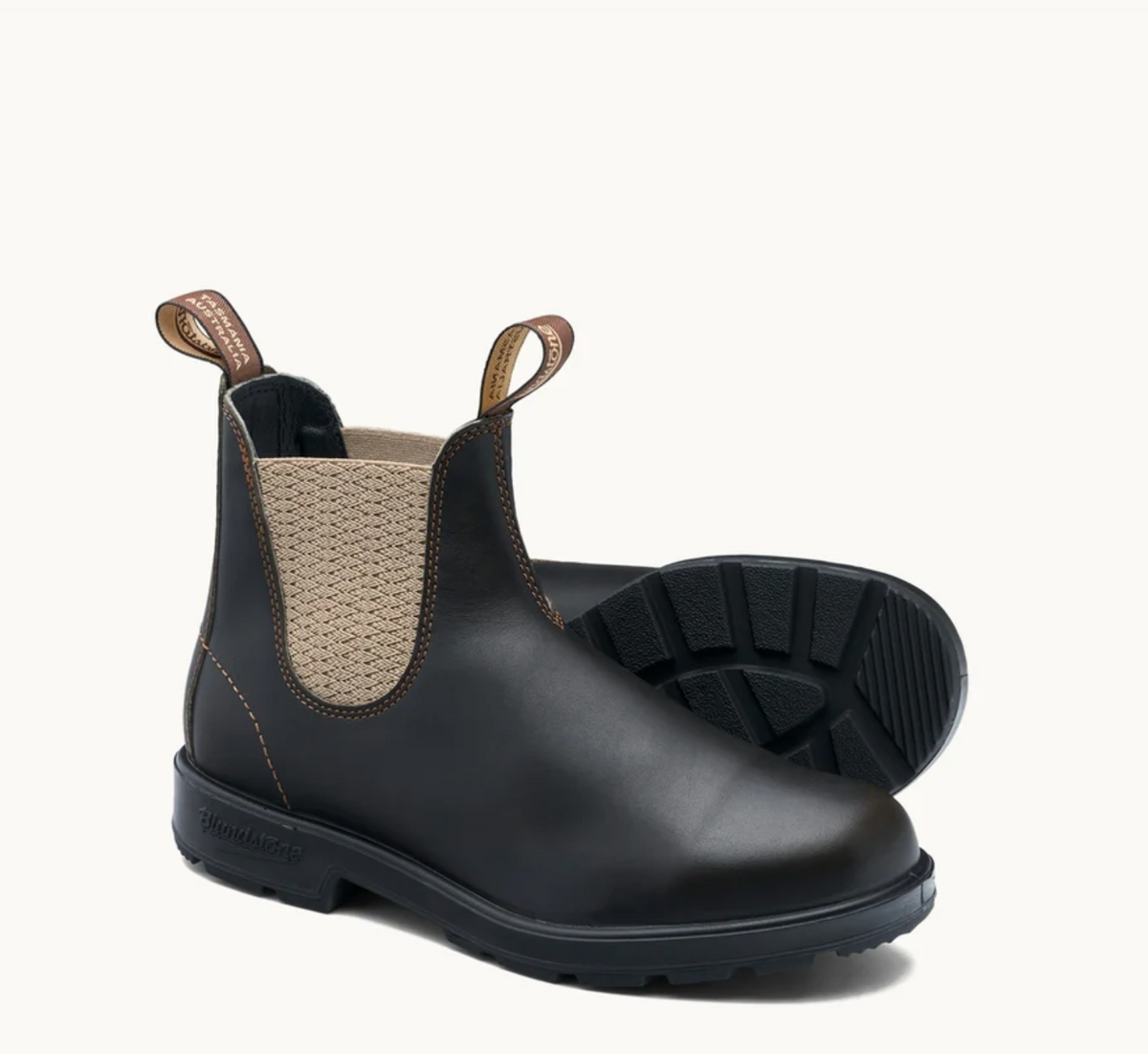 A pair of Blundstone Chelsea boots in black leather, featuring a beige knit elastic side panel and pull tabs, with one boot standing upright and the other lying on its side to show the treaded sole.