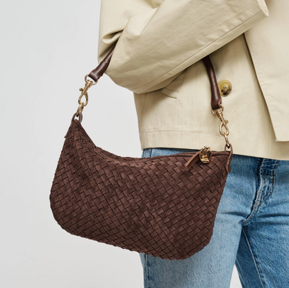 A brown woven leather shoulder bag with gold-tone hardware, shown being worn over the shoulder of a person wearing a beige blazer and blue jeans against a light gray background.