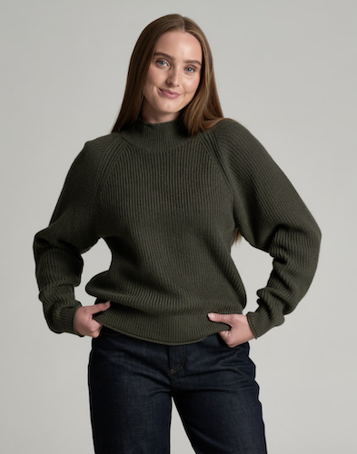 A woman wearing a dark olive green Kuhl Strova ribbed knit mock-neck sweater with balloon sleeves, paired with dark blue jeans, against a solid light gray background.