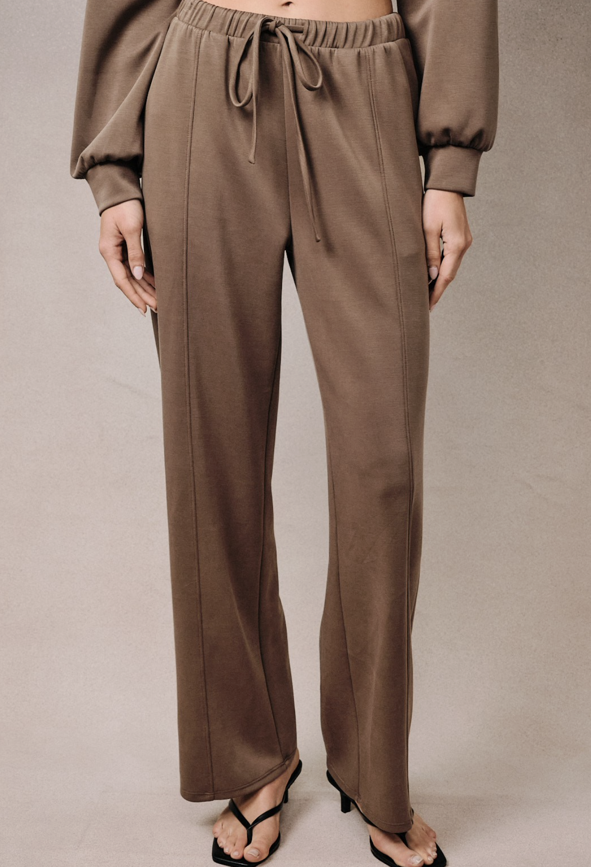 A person wearing loose, wide-leg taupe-brown drawstring pants with a matching long-sleeve top, shown from the chest down against a plain beige studio background.