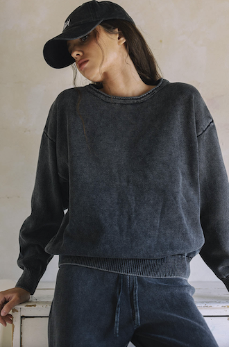 A woman wearing a BL:S BL:S dark washed black crewneck sweatshirt and matching sweatpants, paired with a black baseball cap, leaning against a rustic white wooden bench against a textured beige wall.