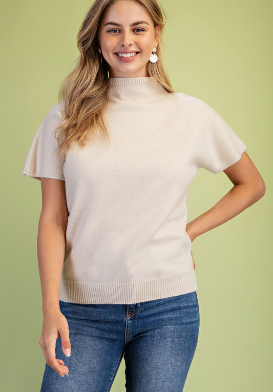 A woman wearing a short-sleeve mock neck knit sweater in cream and blue jeans, standing against a solid light green background.