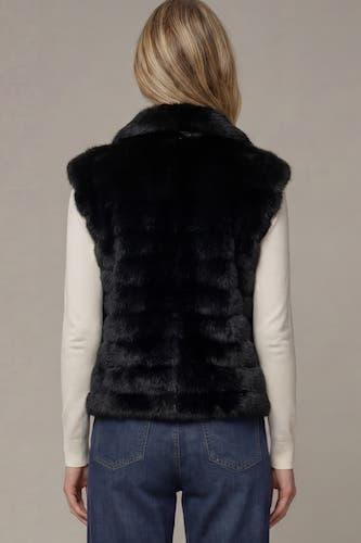 A woman viewed from the back wearing a black faux fur vest over a cream long-sleeve top and blue jeans, against a plain beige studio background.