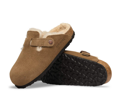 A pair of tan suede Birkenstock clogs with a soft cream shearling lining, shown on a clean white background. One clog is standing upright, displaying the side profile, strap, and the brand name 'BIRKENSTOCK' embossed on the strap.
