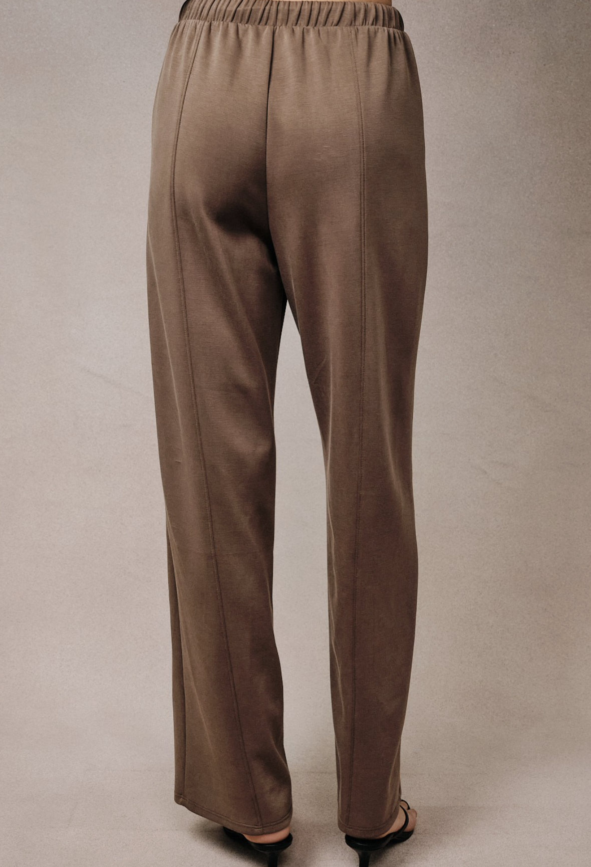A pair of wide-leg taupe pants shown from the back, worn by a model whose feet in black sandals are visible at the bottom, against a neutral beige studio backdrop.