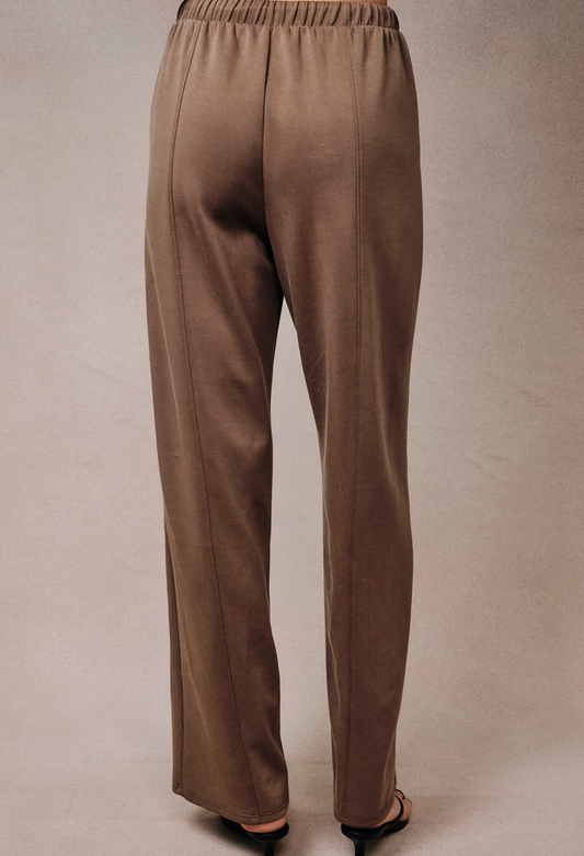 A pair of wide-leg taupe pants shown from the back, worn by a model whose feet in black sandals are visible at the bottom, against a neutral beige studio backdrop.