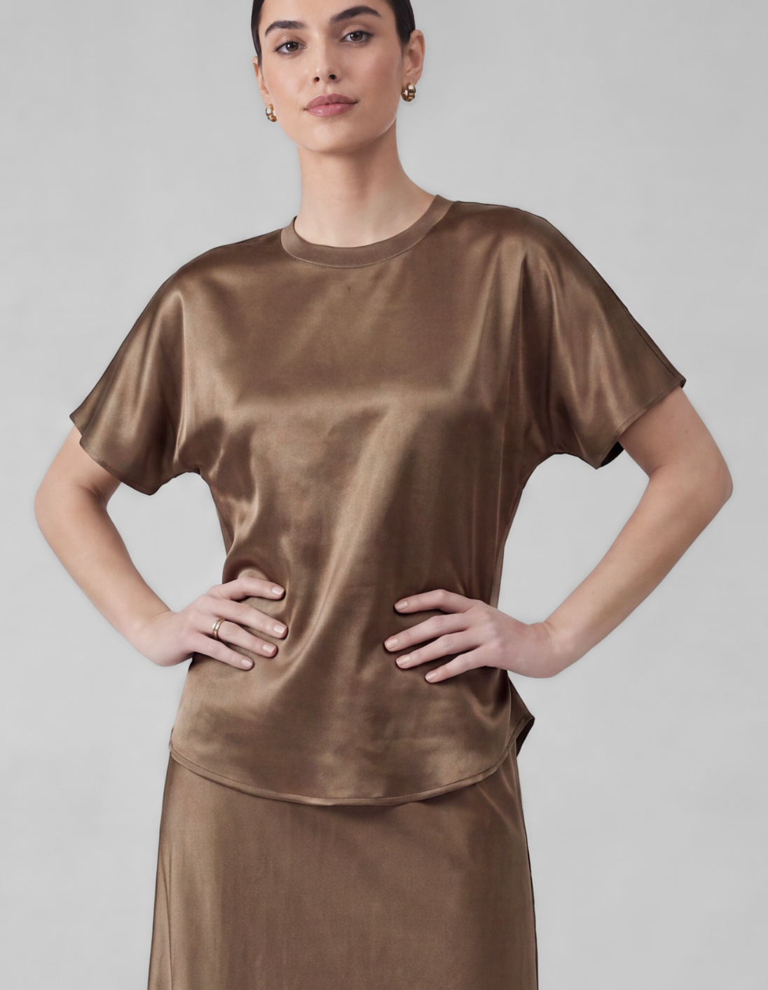 A woman modeling a short-sleeve satin-like top in a warm taupe-brown color, paired with a matching skirt, against a plain light gray studio background.