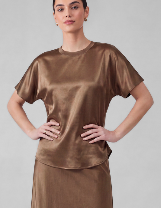 A woman modeling a short-sleeve satin-like top in a warm taupe-brown color, paired with a matching skirt, against a plain light gray studio background.