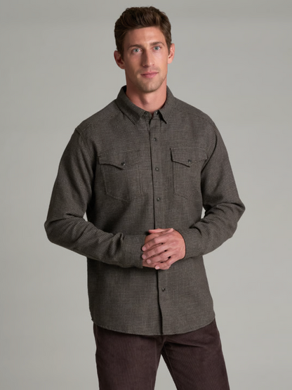 A man wearing a dark gray textured long-sleeve button-down KÜHL Disputr shirt with two chest pockets, paired with dark brown pants, standing against a solid gray studio background.