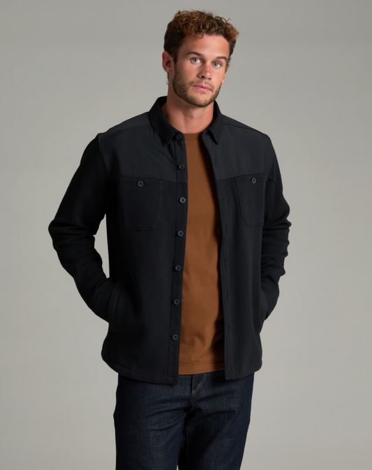 A man wearing a black Kuhl Relentr shirt over a brown t-shirt and dark blue jeans, standing against a solid gray studio background.