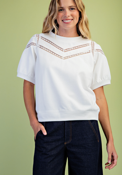 A woman wearing a white short-sleeve crewneck sweatshirt with decorative crochet lace detailing across the chest and shoulders, paired with dark blue jeans, set against a solid light green background.